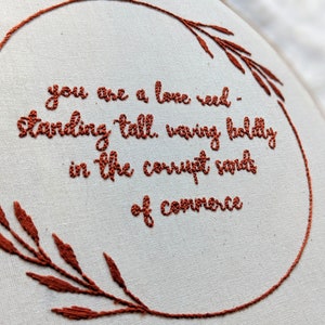 May include: Embroidered hoop art with a white background and brown embroidery. The embroidery reads "you are a lone reed - standing tall, waving boldly in the corrupt sands of commerce".
