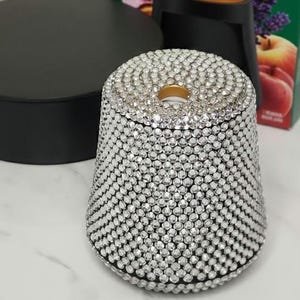 May include: A bedazzled essential mist diffuser with a black base and lid. The diffuser is covered in small, clear rhinestones. A box of Air Wick essential mist diffuser is in the background.