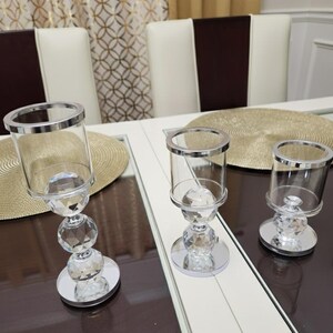 May include: Three crystal candle holders with silver rims and bases. The candle holders are displayed on a white and brown table, with a gold placemat. The background features chairs and patterned curtains.