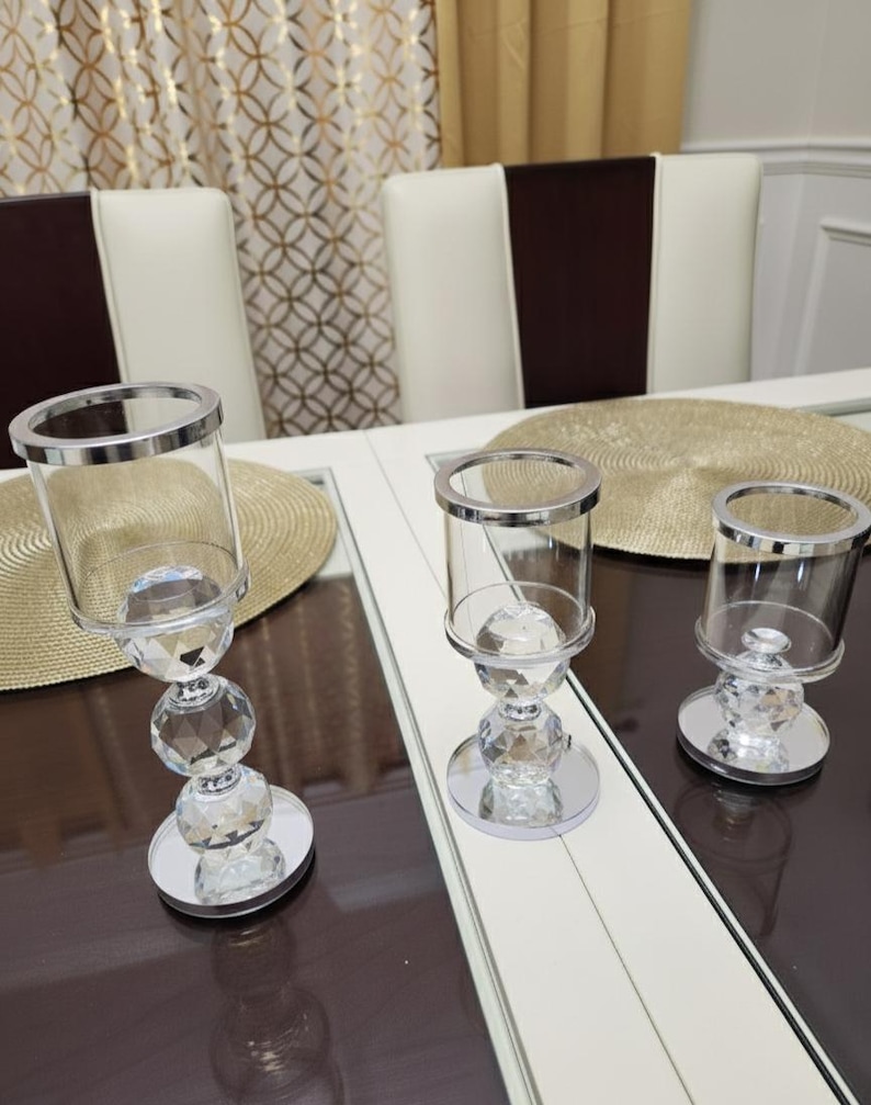 May include: Three crystal candle holders with silver rims and bases. The candle holders are displayed on a white and brown table, with a gold placemat. The background features chairs and patterned curtains.
