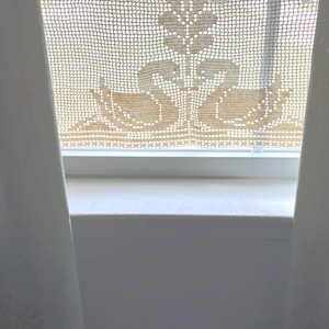 May include: A beige crocheted window curtain featuring a detailed floral and swan design. The curtain is hanging in front of a window. The design includes a central floral element and two swans at the bottom.