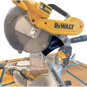May include: A yellow and silver DeWalt mitre saw with a circular blade cutting a piece of wood. The saw has a black handle and a dust collection port. The blade is sharp. The saw is on a wooden surface.