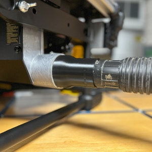 May include: Close-up of a black dust port on a table saw, connected to a black flexible hose.