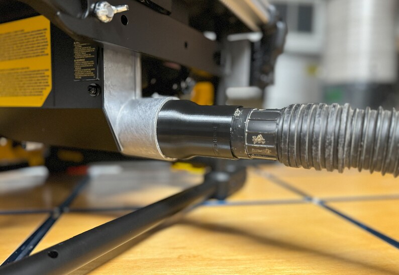 May include: Close-up of a black dust port on a table saw, connected to a black flexible hose.