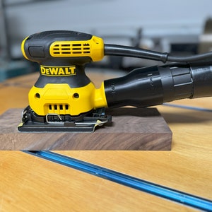 May include: A yellow and black DeWalt electric sander with a black dust collection hose. The sander is sitting on a piece of dark wood.