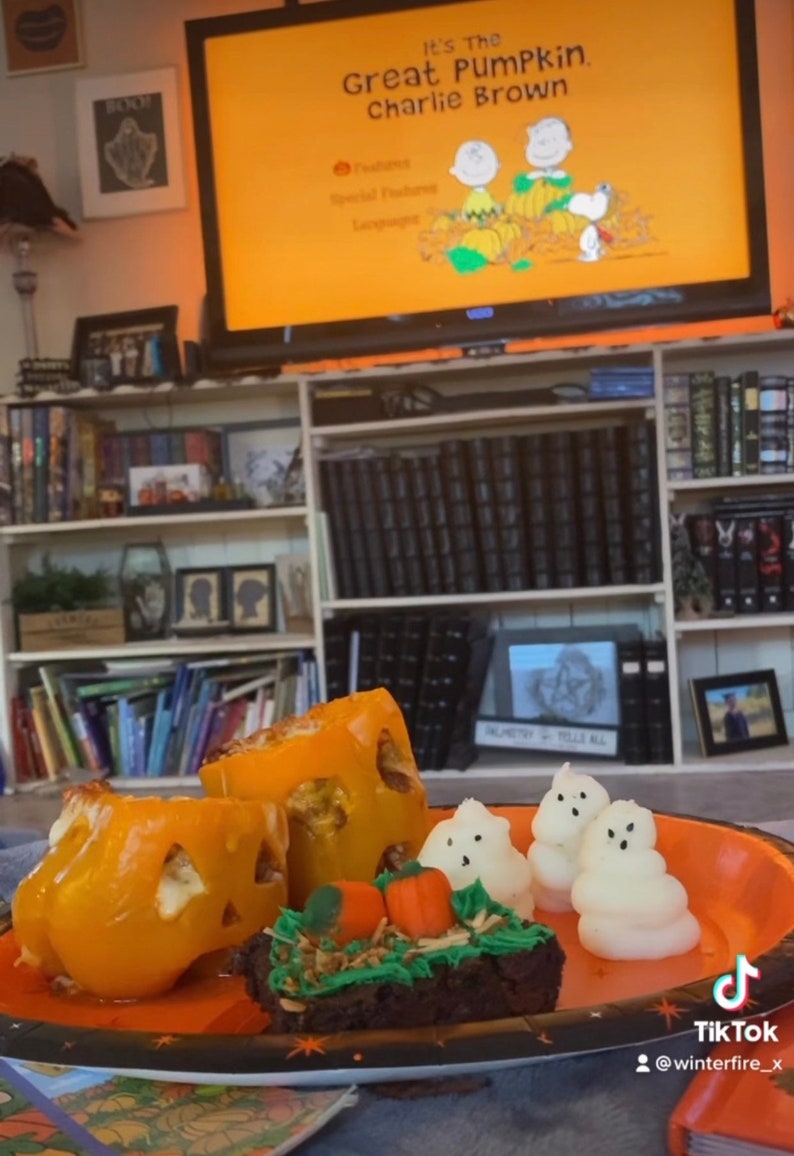 May include: A plate of Halloween-themed treats, including orange bell pepper jack-o'-lanterns, a chocolate brownie with green frosting and candy pumpkins, and white marshmallow ghosts. The plate is on a blue surface with a book and a TV screen in the background. The TV screen shows the title card for the animated special "It's the Great Pumpkin, Charlie Brown."