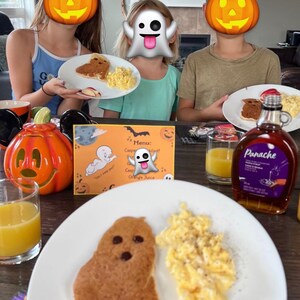 May include: A white plate with a ghost-shaped pancake, scrambled eggs, and apple slices. The pancake has chocolate chips for eyes and a mouth. The plate is on a wooden table with a glass of orange juice, a pumpkin, and a bottle of pancake syrup.