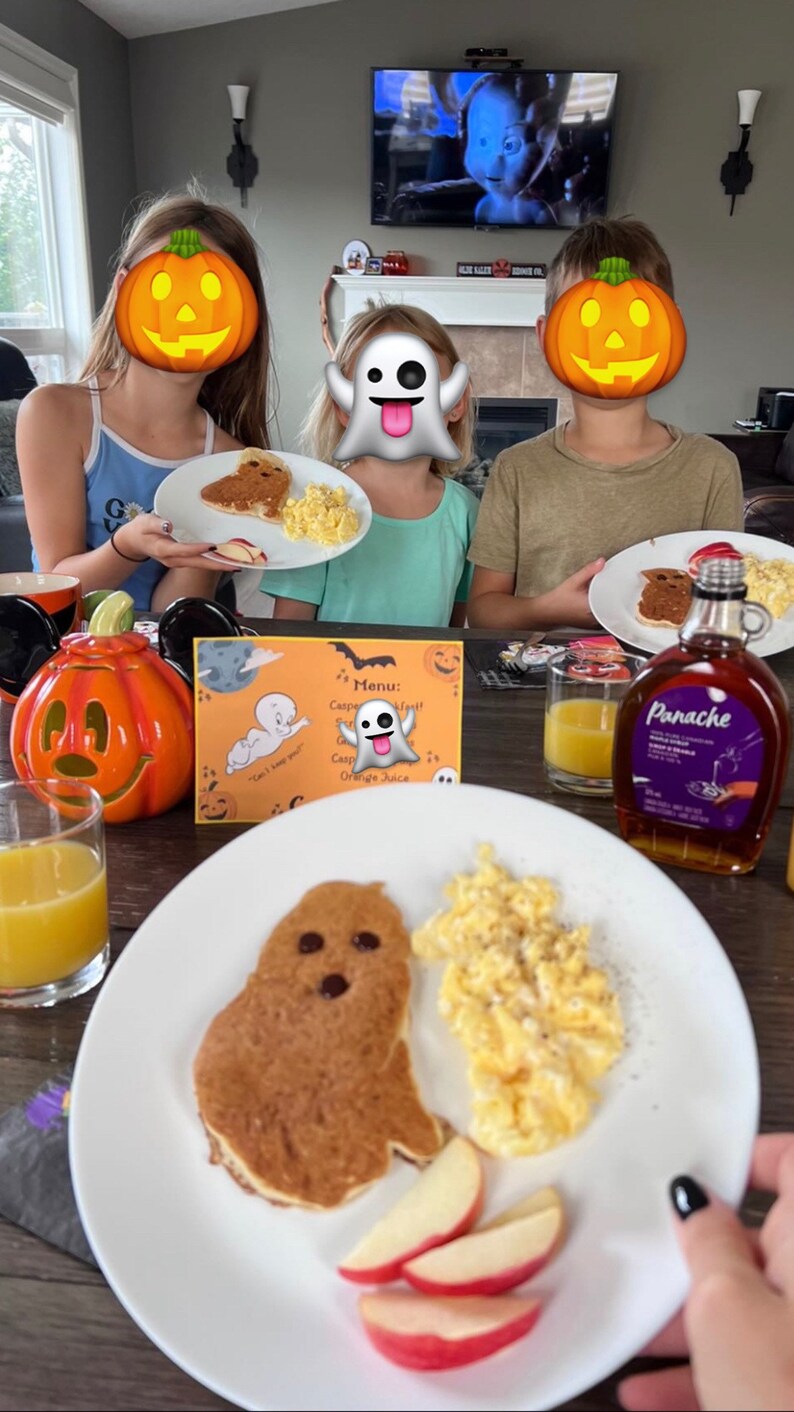 May include: A white plate with a ghost-shaped pancake, scrambled eggs, and apple slices. The pancake has chocolate chips for eyes and a mouth. The plate is on a wooden table with a glass of orange juice, a pumpkin, and a bottle of pancake syrup.