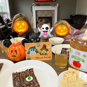 May include: A table setting with a Halloween theme. There are two plates with brownies, one with a green eye on top. There is a glass of apple cider, a bowl of soup, and a pumpkin-shaped lantern. The table is decorated with a Hocus Pocus book and a black cat figurine.