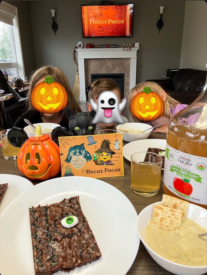 May include: A table setting with a Halloween theme. There are two plates with brownies, one with a green eye on top. There is a glass of apple cider, a bowl of soup, and a pumpkin-shaped lantern. The table is decorated with a Hocus Pocus book and a black cat figurine.