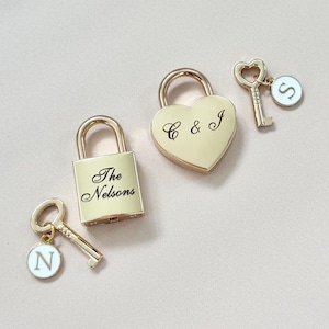 May include: Gold-colored personalized padlocks and keys. One padlock is heart-shaped with "C & J" engraved. Another padlock is rectangular with "The Nelsons" engraved. Each padlock has a matching key with a circular charm.