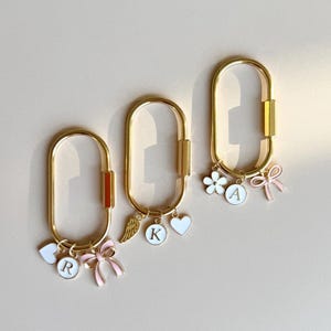 May include: Three gold keyrings with different charms. The charms include a pink bow, a white flower, a white heart, a white angel wing, and white letter charms. The keyrings are on a light brown background.