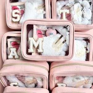 Könnte beinhalten: Rosa Kosmetiktaschen mit transparenten Fenstern, die jeweils weiße, flauschige Artikel, goldene Haarspangen und einen Buchstaben enthalten. Die Buchstaben sind S, T, M und A. Die Taschen sind gestapelt.