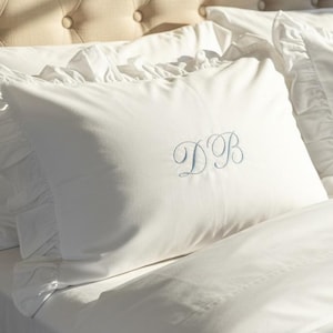 May include: White pillow with blue embroidered initials DB, set on a bed with white linens. The pillow has a ruffled edge and is placed in front of a tufted beige headboard. The bedding is crisp and clean.