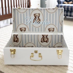 May include: White storage chest with gold hardware, featuring a blue and white striped interior with teddy bear accents. The name "Rhodes" is displayed in light blue script. The chest is open, revealing the interior design.