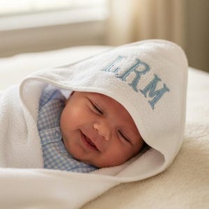 May include: A white hooded baby towel featuring the embroidered blue monogram LRM on the hood. The towel is wrapped around a baby wearing a light blue and white gingham patterned shirt, with the hood pulled up over the baby's head.