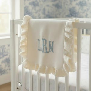 Può includere: Coperta per neonati color crema con bordo a balze, ricamata con le iniziali "LRM" in azzurro. La coperta è drappeggiata su una culla bianca, con una carta da parati a motivi blu e bianchi sullo sfondo.