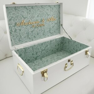 May include: A white rectangular storage chest with gold-toned metal latches and handles. The interior is lined with a sage green fabric featuring a white lace pattern. The lid interior displays the gold text "Mackenzie & Alex 12.12.2026".