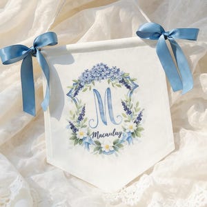 May include: White banner with a floral wreath design in shades of blue and green, featuring the letter "M" and the name "Macaulay". The banner is adorned with light blue satin bows at the top for hanging.