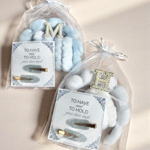 May include: Two gift bags filled with light blue and white plush items, each topped with a jeweled initial. Each bag includes a card that reads "TO HAVE and TO HOLD your hair back" and two gold-toned hair clips.
