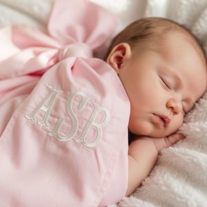 Puede incluir: Un bebé dormido envuelto en una manta rosa claro con un gran lazo. La manta está bordada con las iniciales "ASB" en blanco. El bebé está acostado sobre una manta suave y blanca.