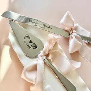 May include: A silver cake knife and server set with a pink ribbon bow. The knife is engraved with "Charlotte & Bradley June 11, 2026". The server is engraved with "C & B 6.17.2026" and a heart.