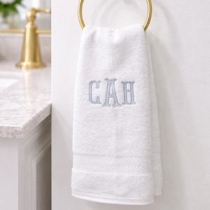 Puede incluir: Una toalla de mano blanca con las iniciales "CAH" bordadas en azul claro, colgada de un aro dorado. El entorno del baño es luminoso y minimalista. La toalla está hecha de un material suave y absorbente.