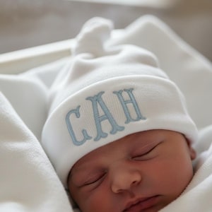Könnte beinhalten: Ein Neugeborenes mit einer weißen Strickmütze mit den hellblau gestickten Initialen "CAH". Das Baby ist in eine weiße Decke gehüllt. Die Mütze hat einen Knoten oben. Die Augen des Babys sind geschlossen.