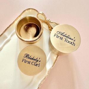 Pode incluir: Duas caixas de lembrança douradas com tampas gravadas. Uma caixa diz "Adeline's First Tooth" e a outra diz "Blakely's First Curl".