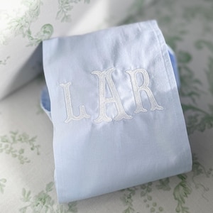 May include: A light blue cloth with the embroidered letters "LAR" in white. The cloth is folded and rests on a surface with a floral pattern in green and white. The image is well-lit, highlighting the details of the embroidery.
