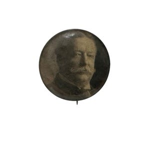 May include: A vintage button featuring a black and white photo of a man with a mustache. The button is round and has a metal pin on the back.