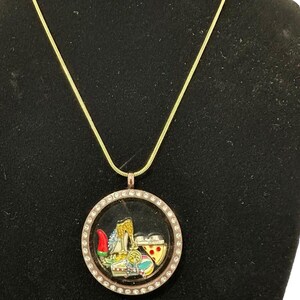 May include: Gold-tone necklace with a round pendant. The pendant features a clear center with various colorful charms, including a high heel, pizza slice, and a red hat. The pendant is surrounded by a ring of small, clear crystals.