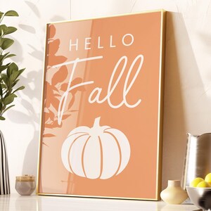 May include: A framed print with a peach background and white text that says "Hello Fall". A white silhouette of a pumpkin is below the text.