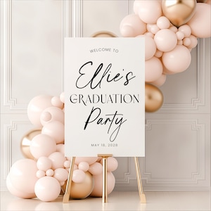 May include: A white graduation party sign on a gold easel, with the text "Welcome to Ellie's Graduation Party" in black script. The date "May 18, 2028" is below. The sign is surrounded by pink and gold balloons.