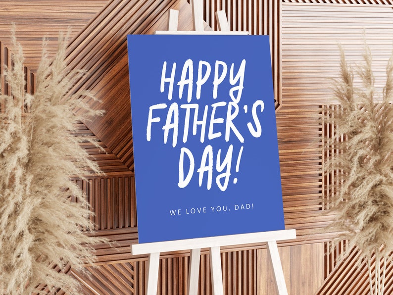 May include: A blue sign on a white easel with the text "HAPPY FATHER'S DAY!" in white, handwritten-style font. Below the main text is the phrase "WE LOVE YOU, DAD!" The background is a wooden wall with decorative panels.