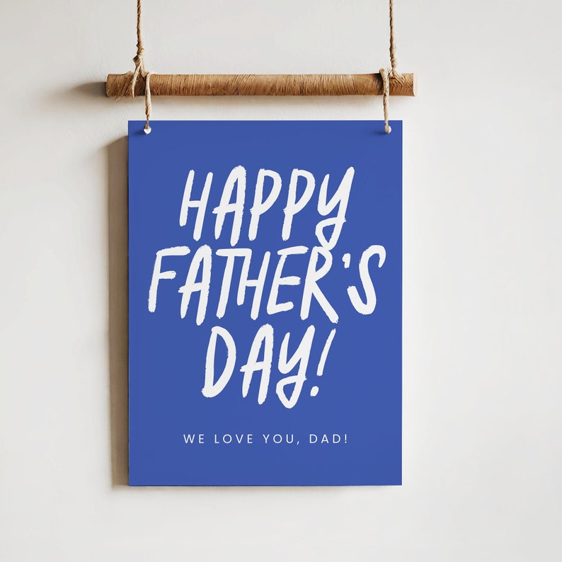 May include: A blue rectangular sign with the words "HAPPY FATHER'S DAY!" in white, handwritten-style lettering. Below, the text "WE LOVE YOU, DAD!" is also in white. The sign is suspended by a wooden dowel and twine.