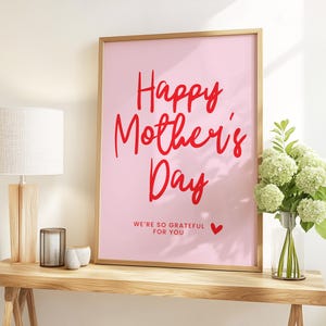 May include: A pink framed print with the text "Happy Mother's Day" in red lettering. The text "We're so grateful for you" is written in smaller red lettering below the main text. A small red heart is to the right of the text.
