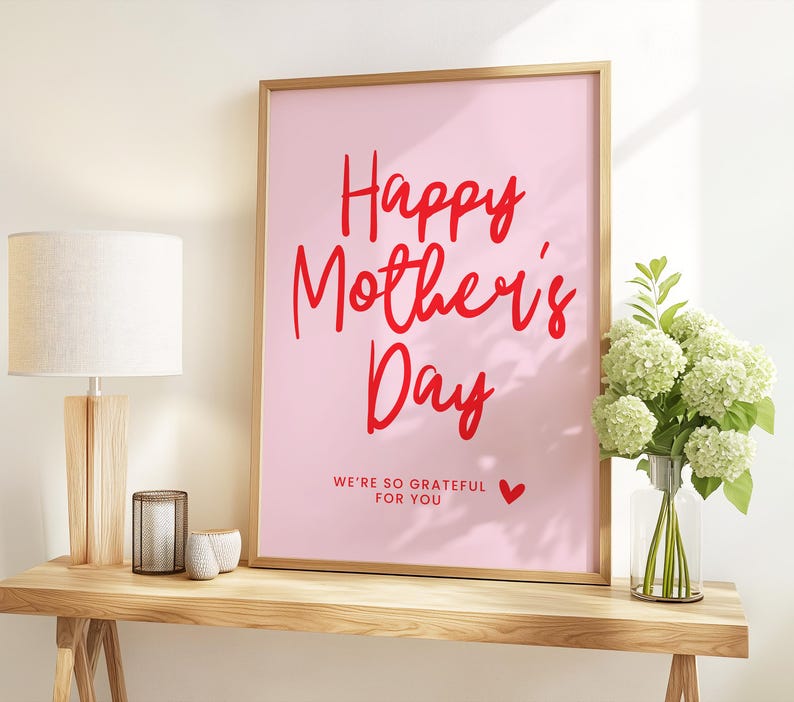 May include: A pink framed print with the text "Happy Mother's Day" in red lettering. The text "We're so grateful for you" is written in smaller red lettering below the main text. A small red heart is to the right of the text.