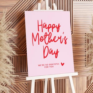 May include: A pink sign with a white easel reads "Happy Mother's Day" in red cursive lettering. Below the greeting, it says "We're so grateful for you" in smaller red letters with a small red heart.