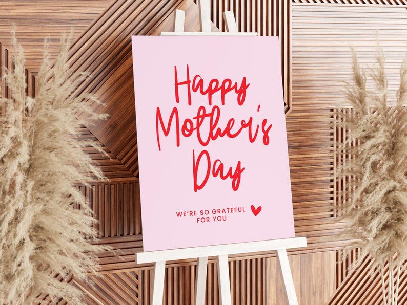 May include: A pink sign with a white easel reads "Happy Mother's Day" in red cursive lettering. Below the greeting, it says "We're so grateful for you" in smaller red letters with a small red heart.