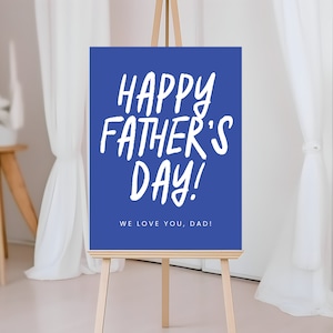 May include: A blue sign on an easel with the text "HAPPY FATHER'S DAY!" in white, handwritten-style font. Below, it reads "WE LOVE YOU, DAD!" The sign is displayed in a bright room with white curtains and a wooden side table.