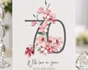 May include: A white greeting card featuring the number 70 in a dark gray font, decorated with layered pink and white floral illustrations. The text reads With love on your 70TH BIRTHDAY in a black script and sans-serif font on textured paper.
