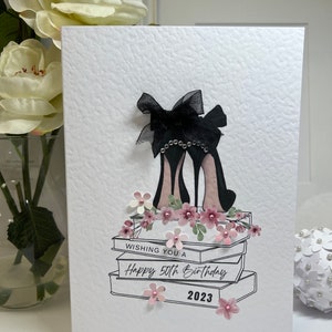 May include: A white card with a black and white illustration of high heels on a stack of books. The card says "Wishing you a Happy 50th Birthday 2023".