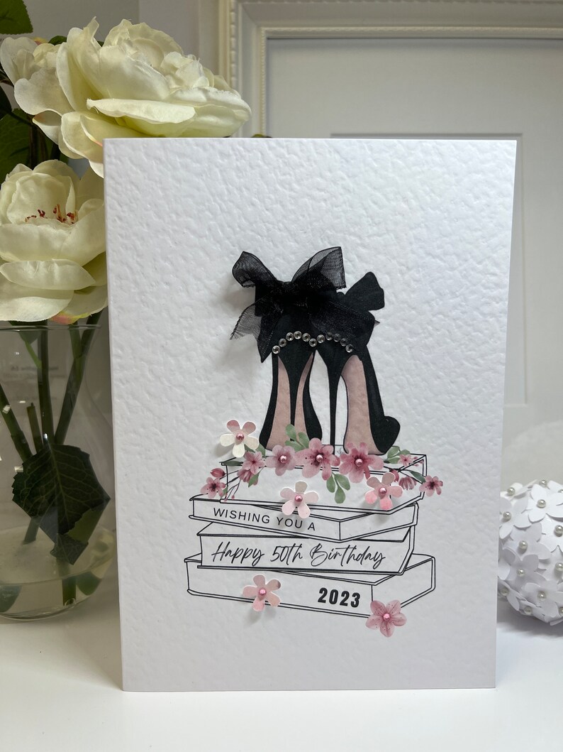 May include: A white card with a black and white illustration of high heels on a stack of books. The card says "Wishing you a Happy 50th Birthday 2023".