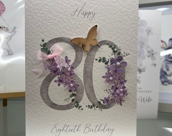 May include: A white card with the words "Happy Eightieth Birthday" written in cursive. The number 80 is decorated with purple flowers and a gold butterfly.