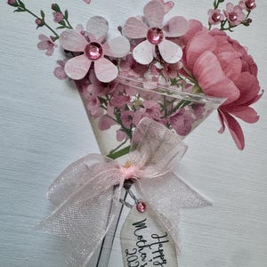 以下が含まれることがあります： 手作りの母の日カード。ピンク色の紙の花束で、それぞれにラインストーンの中心があります。 花束は透明なピンクのリボンで結ばれ、「Happy Mother's Day 2026」と書かれたタグが付いています。 背景は白で、小さなピンクのハートがあります。