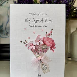 以下が含まれることがあります： 「With Love To A Very Special Mum」のメッセージが書かれた母の日カード。ピンクの花とリボンで飾られたマティーニグラスに花がアレンジされています。小さなタグには「Happy Mother's Day 2023」と書かれています。