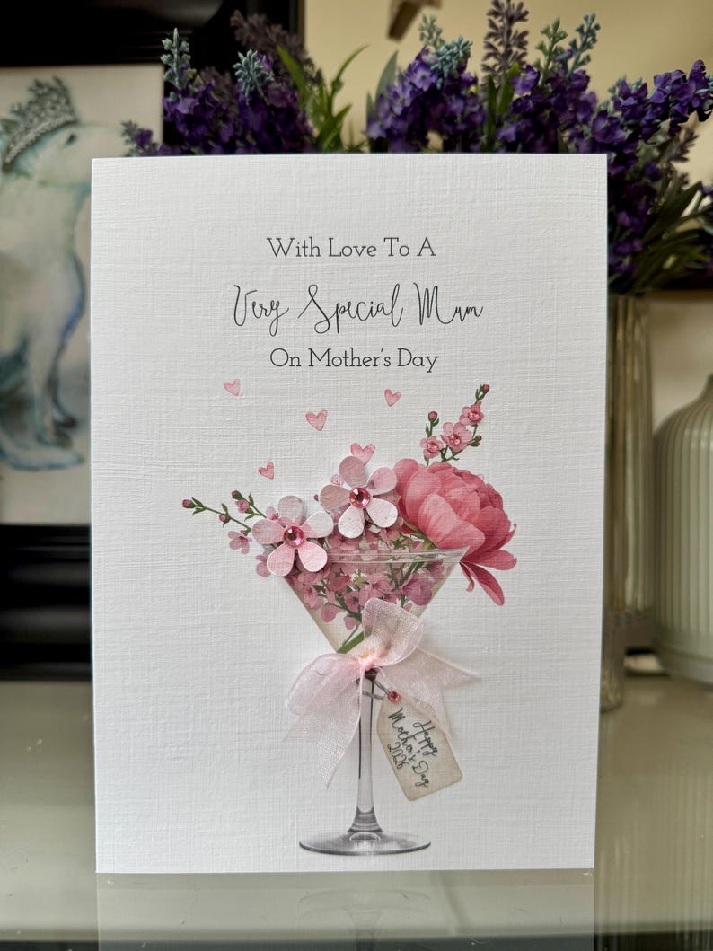 以下が含まれることがあります： 「With Love To A Very Special Mum」のメッセージが書かれた母の日カード。ピンクの花とリボンで飾られたマティーニグラスに花がアレンジされています。小さなタグには「Happy Mother's Day 2023」と書かれています。