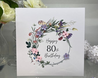 May include: A white card with a floral wreath design. The wreath is made of pink and purple flowers and green leaves. The text "Happy 80th Birthday" is written in black lettering inside the wreath.