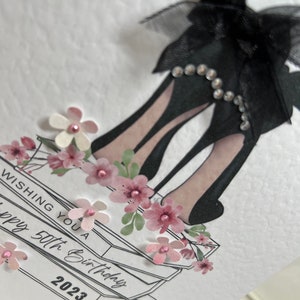 May include: A white card with a black high heel shoe with a black bow and rhinestones. The card has a stack of books with pink flowers and the text "Wishing you a happy 50th Birthday 2023".
